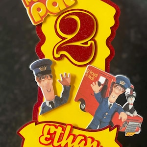 Może przedstawiać: Topper na tort w stylu Postman Pat. Topper zawiera napisy "Postman Pat" i cyfrę "2" w kolorze czerwonym i żółtym. Imię "Ethan" jest również uwzględnione. Obraz zawiera ilustracje Postmana Pata i jego ciężarówki pocztowej.