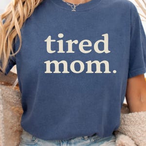 Puede incluir: Una camiseta azul marino con las palabras "tired mom." en blanco. La camiseta de manga corta parece ser de un material suave. La persona lleva pantalones cortos vaqueros y un c&aacute;rdigan color crema. Decoraciones oto&ntilde;ales.