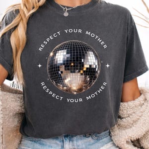 May include: A charcoal gray short-sleeve t-shirt featuring a central graphic of a reflective disco ball. The text "RESPECT YOUR MOTHER" is printed in white capital letters in a circular arc above and below the disco ball, accented by small stars.