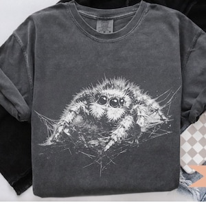 Pu&ograve; includere: T-shirt grigio scuro con un'illustrazione bianca di un ragno saltatore. Il disegno del ragno &egrave; molto dettagliato, mostrando i suoi occhi e il suo corpo. La maglietta &egrave; presentata con le maniche arrotolate ed &egrave; abbinata a jeans e una custodia per telefono. La parola "PEPPER" &egrave; visibile.