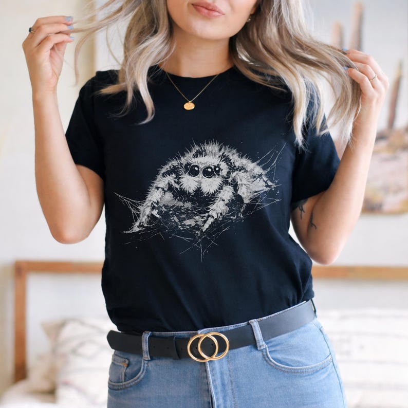 Pu&ograve; includere: T-shirt nero con un'illustrazione dettagliata in scala di grigi di un ragno saltatore. Il ragno &egrave; il punto focale, su uno sfondo a forma di ragnatela. La maglietta ha maniche corte e girocollo, abbinata a jeans blu e una cintura nera.