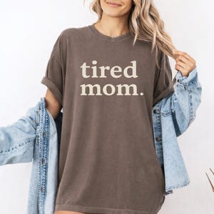 Puede incluir: Una camiseta marr&oacute;n con las palabras "tired mom." en blanco y negrita. La camiseta tiene mangas cortas y un corte holgado. Una chaqueta vaquera azul claro est&aacute; sobre los hombros.