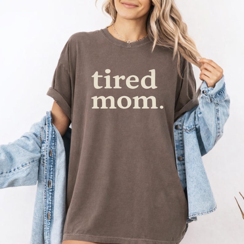 Puede incluir: Una camiseta marr&oacute;n con las palabras "tired mom." en blanco y negrita. La camiseta tiene mangas cortas y un corte holgado. Una chaqueta vaquera azul claro est&aacute; sobre los hombros.