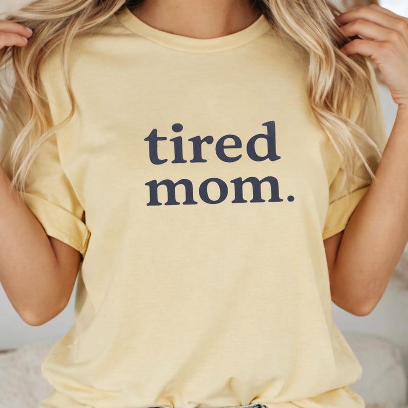 Puede incluir: Camiseta amarilla clara con la frase "tired mom." en azul marino. La camiseta tiene mangas cortas y cuello redondo. El texto est&aacute; en una fuente sencilla sin serifa.