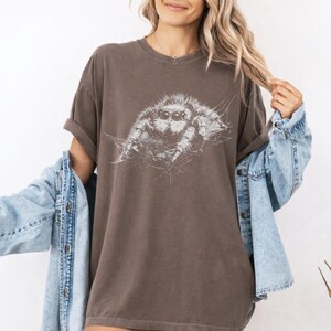 Pu&ograve; includere: T-shirt marrone con un'illustrazione bianca di un ragno saltatore. Il ragno &egrave; il punto focale, con i suoi otto occhi e il suo corpo peloso. La maglietta ha maniche corte e una vestibilit&agrave; ampia. Il colore &egrave; un marrone tenue.