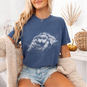 Pu&ograve; includere: Una maglietta blu navy con un'illustrazione bianca di un ragno saltatore. Il ragno &egrave; raffigurato con i suoi occhi e il suo corpo, su una ragnatela. La maglietta ha maniche corte e girocollo.