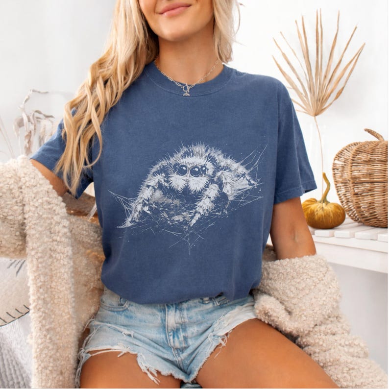 Pu&ograve; includere: Una maglietta blu navy con un'illustrazione bianca di un ragno saltatore. Il ragno &egrave; raffigurato con i suoi occhi e il suo corpo, su una ragnatela. La maglietta ha maniche corte e girocollo.