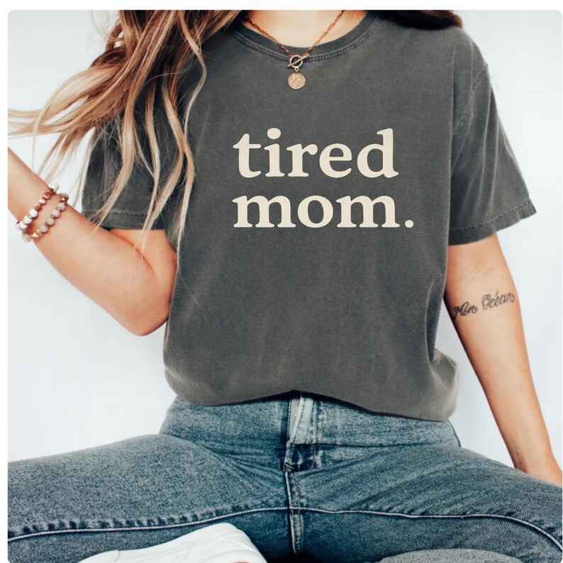 Puede incluir: Camiseta gris oscuro con las palabras "tired mom." en letras blancas. La camiseta se usa con vaqueros azules. La modelo lleva un collar y una pulsera.