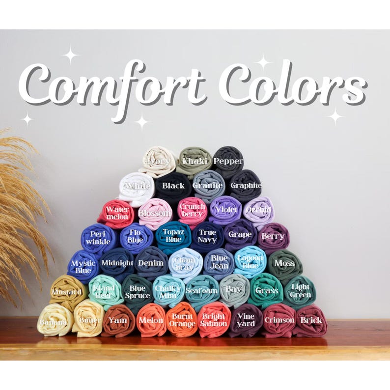 Pu&ograve; includere: Un'esposizione di t-shirt Comfort Colors arrotolate in vari colori, tra cui bianco, nero e blu. L'immagine presenta una disposizione a piramide delle magliette, con nomi di colori come "Watermelon" e "Berry".