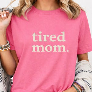 Puede incluir: Camiseta rosa con la frase "tired mom." en beige claro. La camiseta tiene mangas cortas y cuello redondo. El texto est&aacute; centrado en la parte delantera de la prenda.
