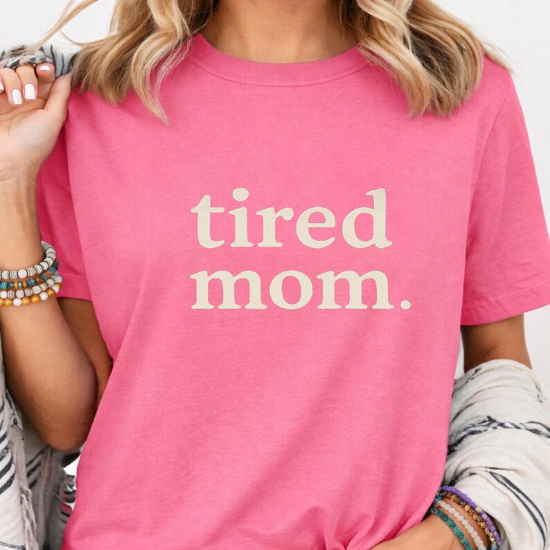 Puede incluir: Camiseta rosa con la frase "tired mom." en beige claro. La camiseta tiene mangas cortas y cuello redondo. El texto est&aacute; centrado en la parte delantera de la prenda.