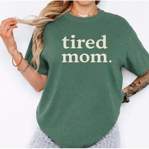 Puede incluir: Una camiseta verde con las palabras "tired mom." impresas en crema. La camiseta tiene mangas cortas y cuello redondo. La imagen muestra la camiseta en una persona.