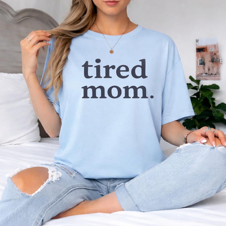 Puede incluir: Camiseta azul claro con la frase "tired mom." en gris oscuro. La camiseta se combina con vaqueros azules claros. La imagen transmite un estilo relajado y c&oacute;modo.