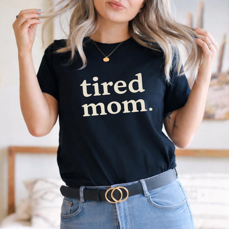 Puede incluir: Camiseta negra con las palabras "tired mom." en una fuente blanca y en negrita. La camiseta se combina con vaqueros azules y un cintur&oacute;n negro con una hebilla dorada de doble c&iacute;rculo. La modelo lleva un collar de oro.