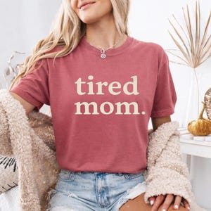 Puede incluir: Camiseta de color rosa polvoriento con las palabras "tired mom." impresas en crema. La camiseta tiene mangas cortas y cuello redondo. La modelo lleva pantalones cortos vaqueros azules y una rebeca crema. La tela es suave.
