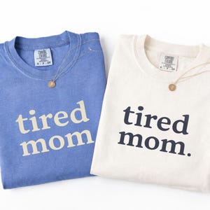 Puede incluir: Dos camisetas Comfort Colors, una azul claro y otra blanquecina, cada una con la frase "tired mom" impresa en la parte delantera. Un collar dorado con un colgante redondo descansa sobre cada camisa. Las camisas est&aacute;n dobladas.