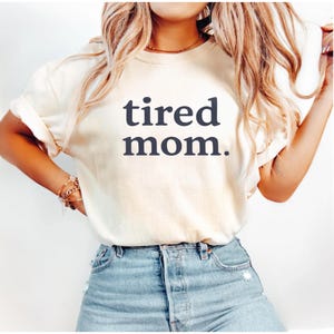 Puede incluir: Camiseta color crema con la frase "tired mom." en azul oscuro. La camiseta tiene mangas cortas y se combina con vaqueros azules. El estilo es informal y c&oacute;modo.