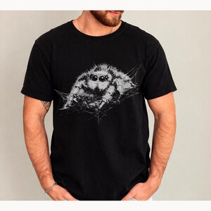 Pu&ograve; includere: T-shirt nero con un'illustrazione dettagliata in scala di grigi di un ragno saltatore. Il ragno ha grandi occhi e intricati dettagli di ragnatela, che contrastano con il tessuto scuro. La maglietta &egrave; un classico girocollo.