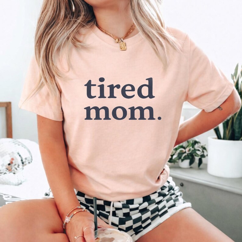 Puede incluir: Una camiseta color melocot&oacute;n con la frase "tired mom." en azul marino. La camiseta tiene mangas cortas y cuello redondo. La persona lleva un pantal&oacute;n corto a cuadros blanco y negro y un collar dorado.