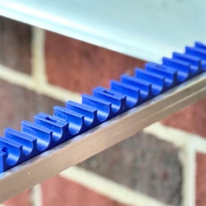 May include: A blue plastic tool holder with multiple slots, designed to hold tools. The holder is mounted on a metal bar, with a brick wall in the background. The tool holder is a vibrant blue color.