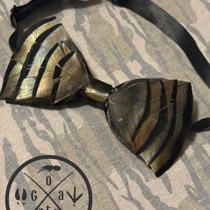 May include: A handmade bow tie featuring a unique feather design with black and gold stripes. The bow tie is attached to a black adjustable strap. The background is a camouflage pattern. The logo "GOAT Outdoors" is visible.