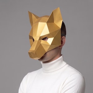 May include: A gold geometric fox mask. The mask is made of folded paper or cardstock, with sharp angles and a detailed design. The person is wearing a white roll neck. The background is a solid grey.