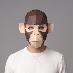 May include: A brown and tan geometric monkey mask. The mask is made of folded paper and covers the face, with eye holes and ear details. The person is wearing a white sweater.