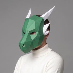 May include: A green geometric dragon mask with white horns and ear accents. The mask is made of paper and worn by a person wearing a white turtleneck sweater. The mask has a polygonal design.
