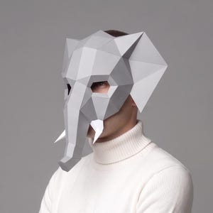 Può includere: Una maschera geometrica grigia a forma di elefante con zanne appuntite. La maschera è realizzata in carta o cartoncino piegato e indossata da una persona che indossa un dolcevita bianco. La maschera copre il viso e la testa.