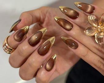 May include: Brown and gold stiletto-shaped nail design. The nails have a glossy, brown base with a gold metallic tip. A gold ring and a gold flower-shaped ring are also visible.