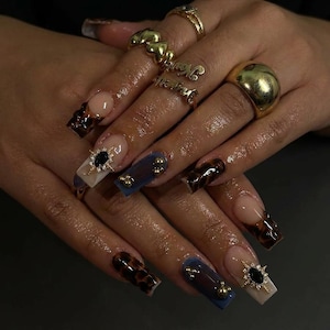 May include: Close-up of a hand with long, manicured nails. The nails feature a variety of designs, including a tortoiseshell pattern, solid blue, and clear with gold accents. Several gold rings adorn the fingers, adding to the overall aesthetic.