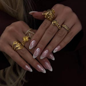 May include: Close-up of hands adorned with multiple gold rings, including a floral design, a geometric pattern, and a lion head. The nails are long, oval-shaped, and painted with a pearlescent pink polish and white tips, creating a French manicure.