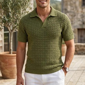 May include: A man wearing a short-sleeved olive green polo shirt and white shorts. The polo shirt has a textured knit pattern and a collared neckline. The shorts are a light color. The man is standing outside on a wooden deck.