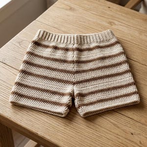 Puede incluir: Pantalones cortos de crochet hechos a mano en color crema con rayas horizontales marrones. Los pantalones cortos están hechos de hilo suave y tienen un ajuste cómodo y relajado. Perfectos para el clima cálido, estos pantalones cortos son una adición elegante y única a cualquier guardarropa.