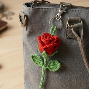 May include: A crochet rose keyring featuring a vibrant red flower head with green leaves and a stem. The rose is attached to a silver-toned metal lobster clasp and ring, which is clipped onto the side of a grey canvas handbag with leather accents.