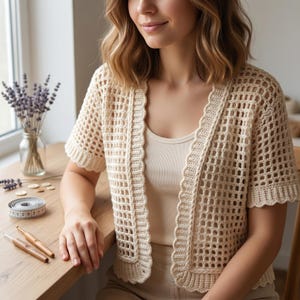 May include: Cream-colored, open-weave crochet cardigan with short sleeves and a scalloped edge. The cardigan is worn over a neutral-toned tank top. A measuring tape, crochet hooks, and a vase of lavender are on a wooden table.
