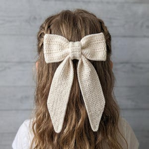 Pode incluir: Um acessório de cabelo em forma de laço, feito de crochê creme. O laço é grande e apresenta um formato de laço clássico com caudas longas e esvoaçantes. O acessório de cabelo é preso ao cabelo, e o fundo é uma parede de painéis de madeira clara.