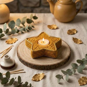 May include: A mustard yellow star-shaped crochet candle holder with a metallic gold trim edge. The holder contains a single lit white tea light candle. The item is positioned on a circular wooden slice, surrounded by scattered gold leaves and eucalyptus.