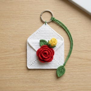 May include: A white crocheted envelope keyring featuring a large red rose and a small yellow rosebud with green leaves attached to the front. A long green crocheted stem with a single leaf hangs from the silver metal ring at the top.