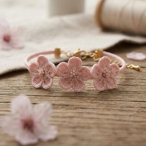 Op de afbeelding: Een delicaat roze armband met drie gehaakte bloemhangers. De bloemen hebben kralen in het midden en zijn bevestigd aan een dun, roze koord met een goudkleurige sluiting. De armband ligt op een houten oppervlak.