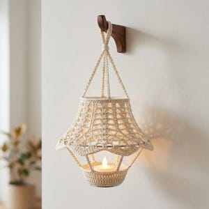 Pode incluir: Uma lanterna de crochê creme com uma vela dentro, pendurada em um gancho de parede de madeira escura. A lanterna tem uma sombra em forma de sino e uma base redonda, com um padrão de tecido aberto decorativo. A vela emite um brilho quente.