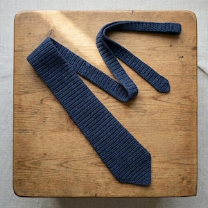 Puede incluir: Una corbata de color azul oscuro confeccionada con un patrón de punto de ganchillo denso y texturizado. La corbata presenta un extremo puntiagudo y una banda larga y estrecha que se dobla sobre sí misma, mostrando una construcción uniforme.