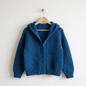 May include: A blue hooded sweater with a ribbed hem and cuffs hangs on a wooden hanger. The sweater is made of a thick, textured knit fabric. The hood is large and the sweater has a buttonless front.