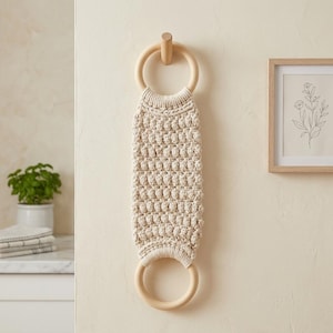 Op de afbeelding: Een crèmekleurige macramé handdoekhouder met twee houten ringen. Het centrale gedeelte heeft een textuur, geweven ontwerp. De houder is gemonteerd op een lichtgekleurde muur, met een ingelijste botanische print op de achtergrond. Een kleine plant in een witte pot is ook zichtbaar.