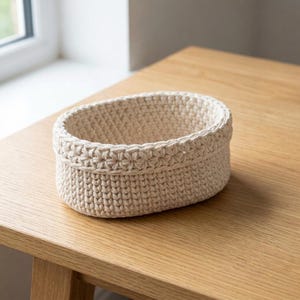May include: A beige, oval-shaped crochet basket. The basket is made of thick yarn and has a textured appearance. The rim features a decorative stitch pattern. The basket sits on a light-colored wooden surface.