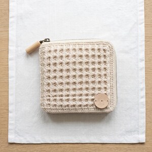Può includere: Un portafoglio beige all'uncinetto con una trama a forma di waffle, un tirazip in legno e un piccolo dettaglio in pelle. Il portafoglio quadrato è chiuso con una cerniera e poggia su una superficie di lino bianco.