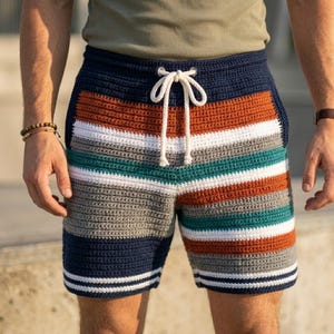 May include: Crocheted shorts with a striped pattern in navy blue, orange, teal, white, and gray. The shorts have a drawstring waist and white stripes at the hem. The shorts are handmade and suitable for casual wear.