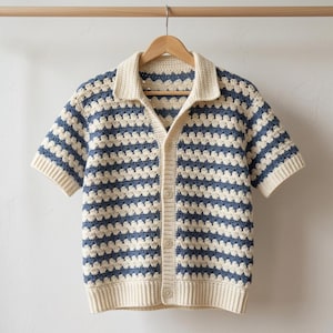 Peut inclure: Une chemise en crochet à manches courtes, rayée crème et bleue, avec un col et une fermeture boutonnée. La chemise est suspendue à un cintre en bois. Le motif au crochet crée un design texturé et ajouré. Mesures en centimètres.