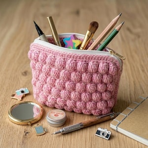 Può includere: Una pochette all'uncinetto rosa con un motivo testurizzato a punto nocciolina e una chiusura a zip bianca. La pochette contiene vari strumenti di scrittura, un pennello per il trucco e una gomma a forma di stella. Vicino ci sono uno specchietto e articoli di cancelleria.