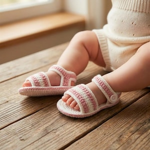 Könnte beinhalten: Handgefertigte Babysandalen in rosa und weißen Streifen. Die Häkelsandalen haben einen Riemen, der mit einem Knopf befestigt wird. Die Sandalen befinden sich auf einer Holzoberfläche.